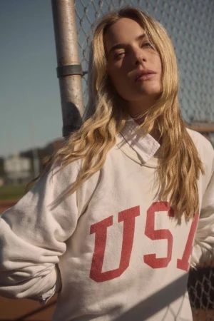 Blonde woman wearing white USA sweatshirt leaning against chain-link fence outdoors in sunlight
