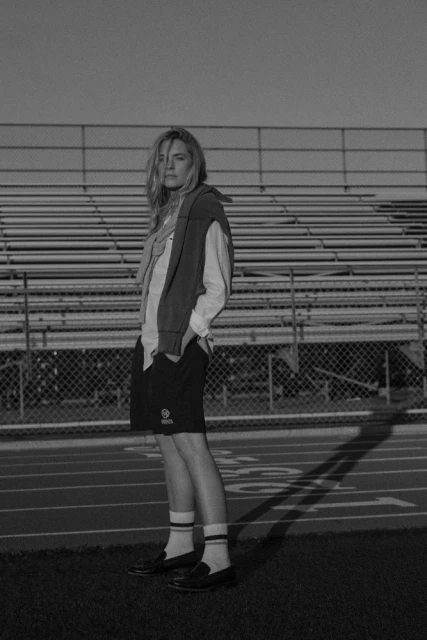 Monochrome portrait of a woman standing on a track field, wearing a sweater, shorts, striped socks, and loafers.