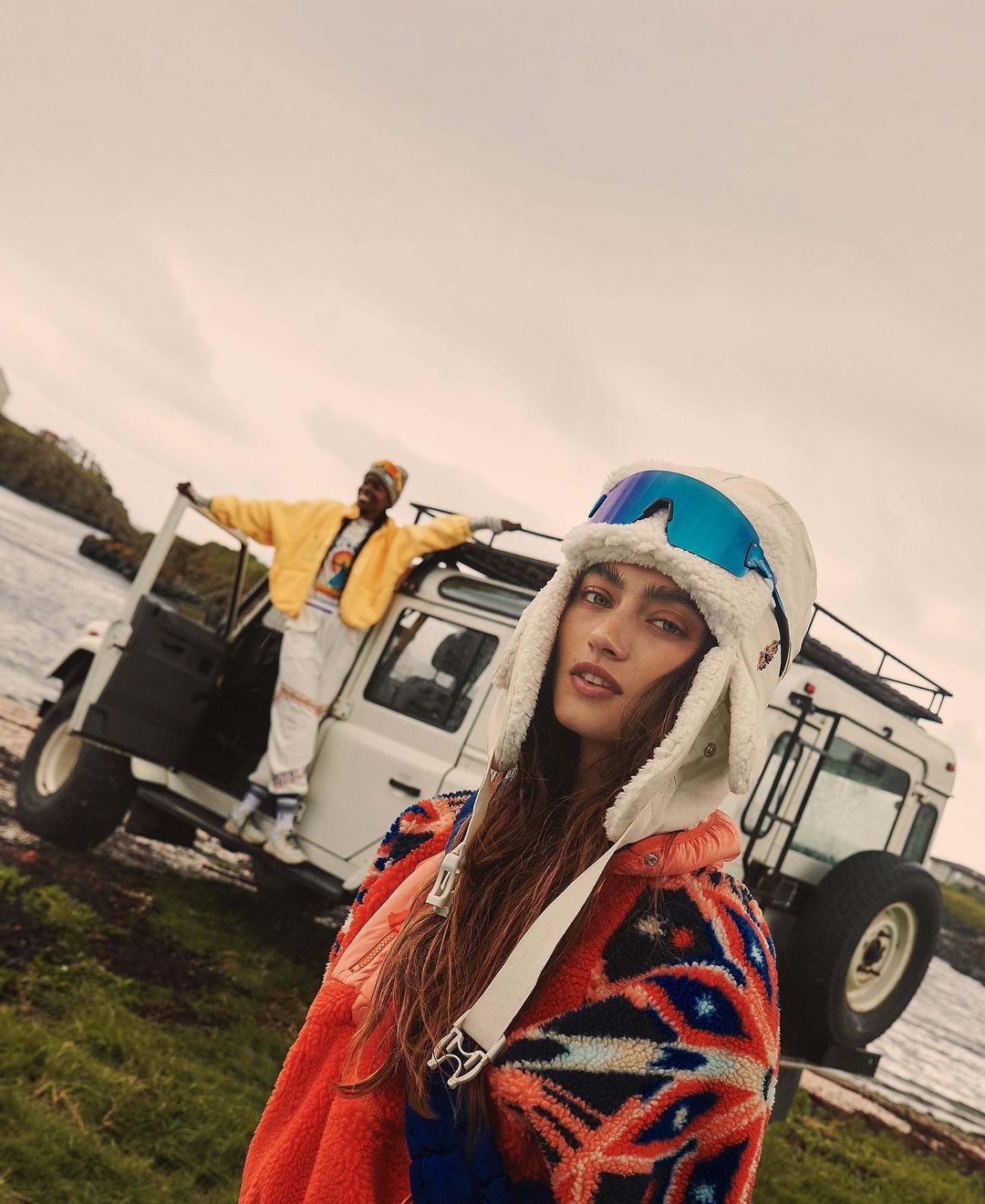 A woman in a colorful patterned jacket and white hat stands in front of an SUV. A man leans out of the vehicle near the ocean.