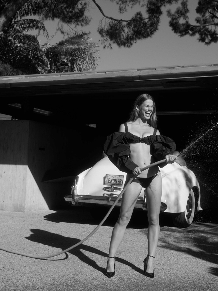 Black and white photograph of a woman holding a garden hose in front of a vintage car and garage on a sunny day.