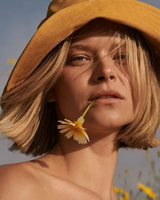 Close-up of a young woman with blonde bob wearing a hat, holding a yellow flower in her mouth against a blue sky and field of flowers.