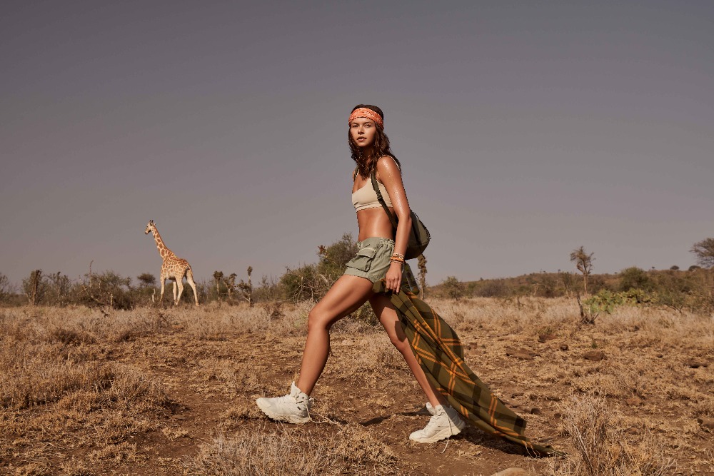 Woman in sportswear walks across dry grassland in sun; giraffe stands in background under clear sky, casual outdoor fashion photo.