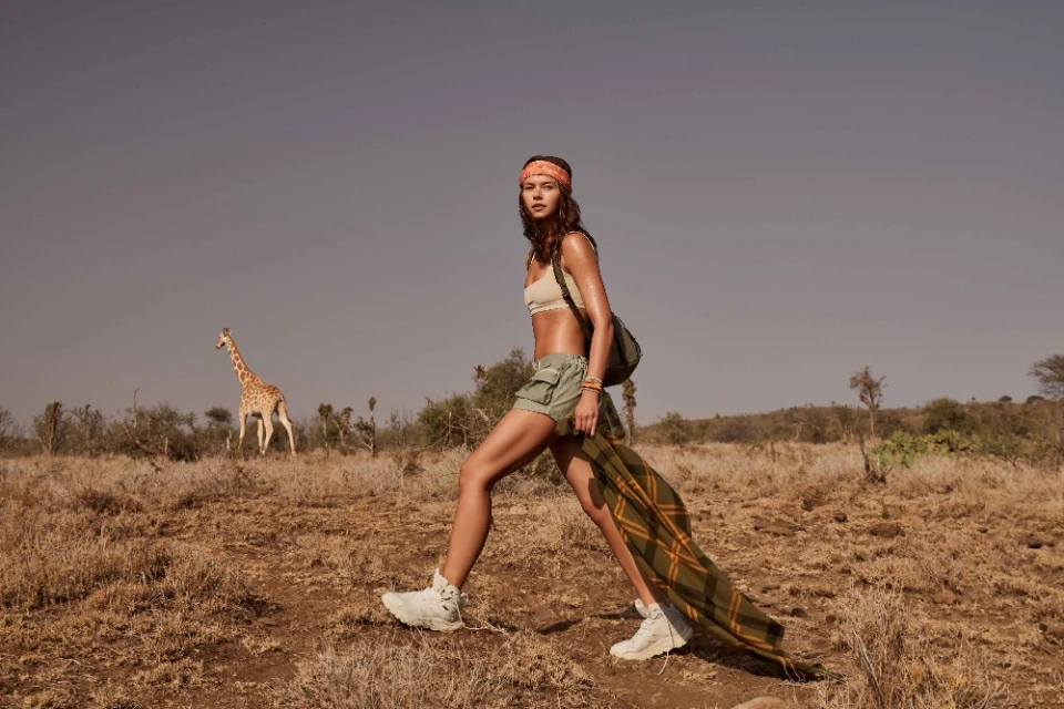 Woman in sportswear walks across dry grassland in sun; giraffe stands in background under clear sky, casual outdoor fashion photo.