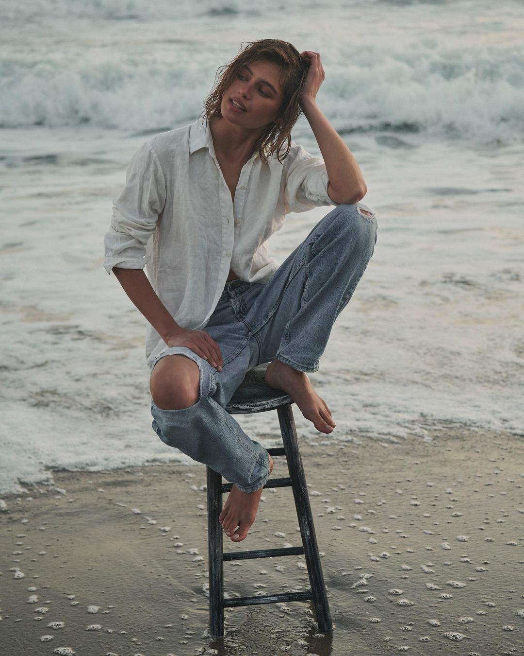 Woman in jeans and a white shirt sits on a black stool at the beach, with waves in the background and sand in the foreground.