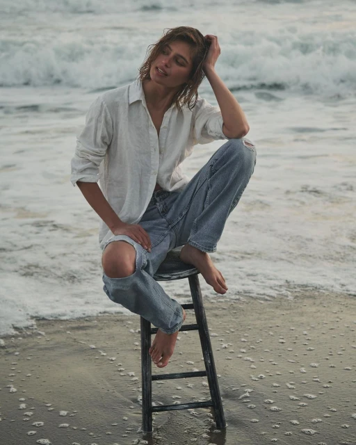 Woman in jeans and a white shirt sits on a black stool at the beach, with waves in the background and sand in the foreground.