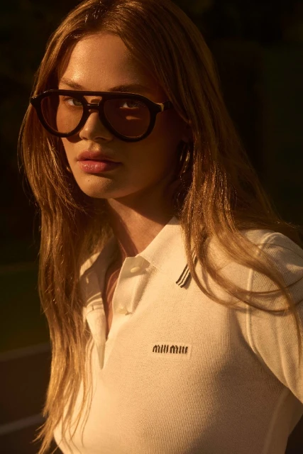 Blonde woman wears stylish aviator sunglasses and a white polo shirt with logo, posing outdoors in diffused natural sunlight.