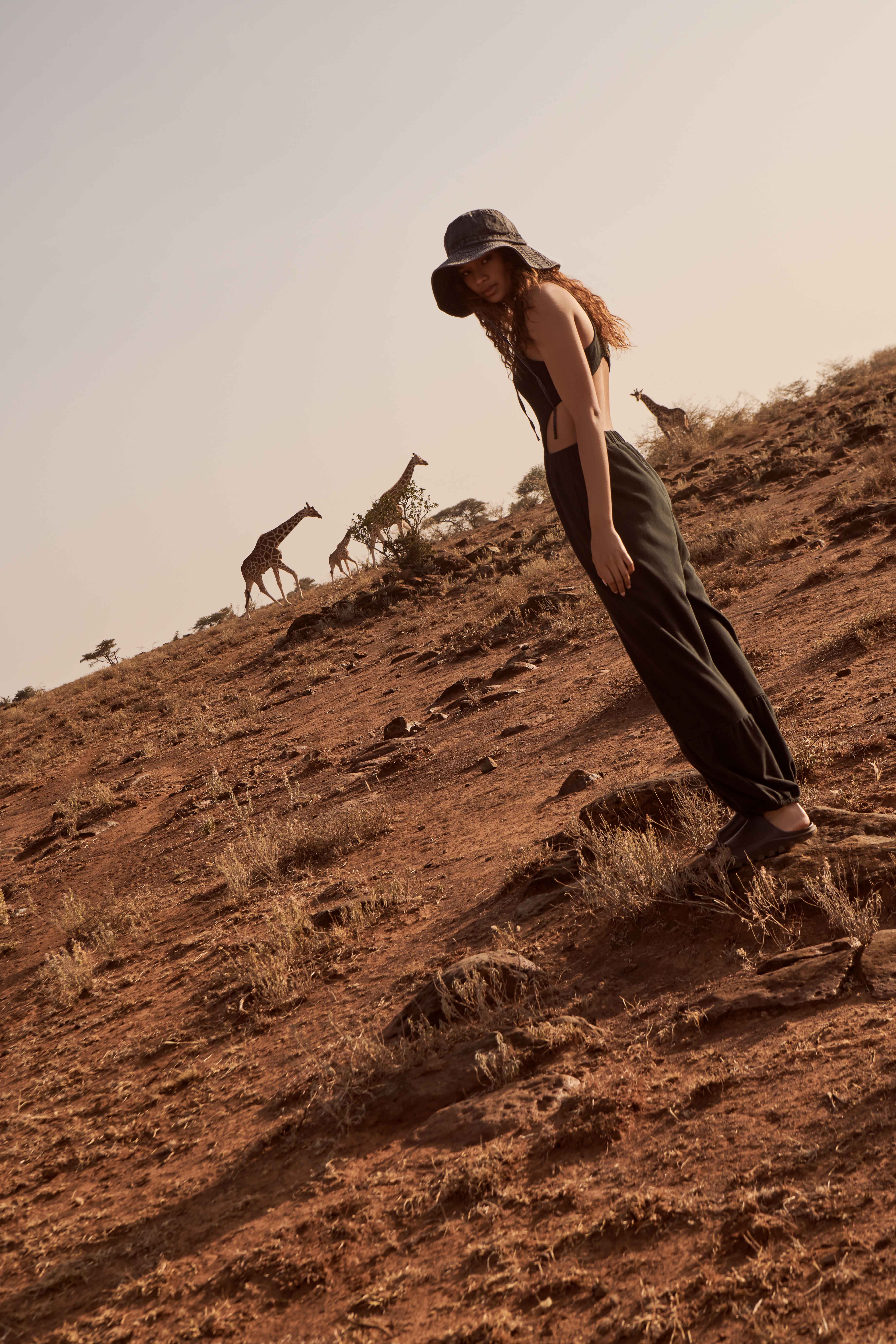 Woman wearing hat and black clothing stands on hillside with giraffes in background, bathed in golden light in an outdoor setting.
