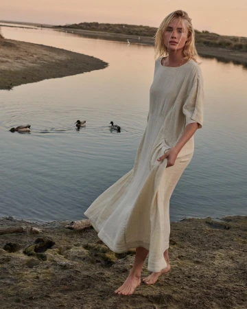 Adult woman in a linen dress poses barefoot near water at dusk. Ducks swim on the water in the soft light of the evening.