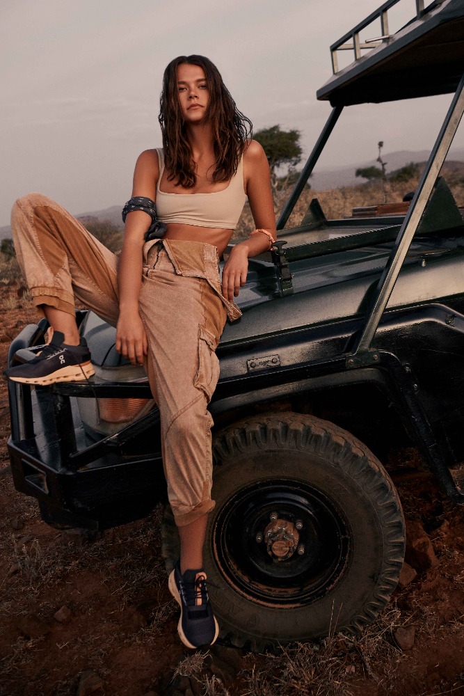 Brunette woman in tan pants and tank top poses on a black vehicle outdoors in a rural setting with dry grass and distant trees.