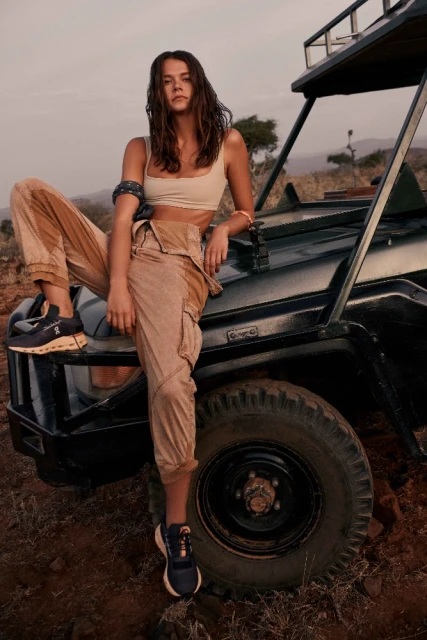 Brunette woman in tan pants and tank top poses on a black vehicle outdoors in a rural setting with dry grass and distant trees.
