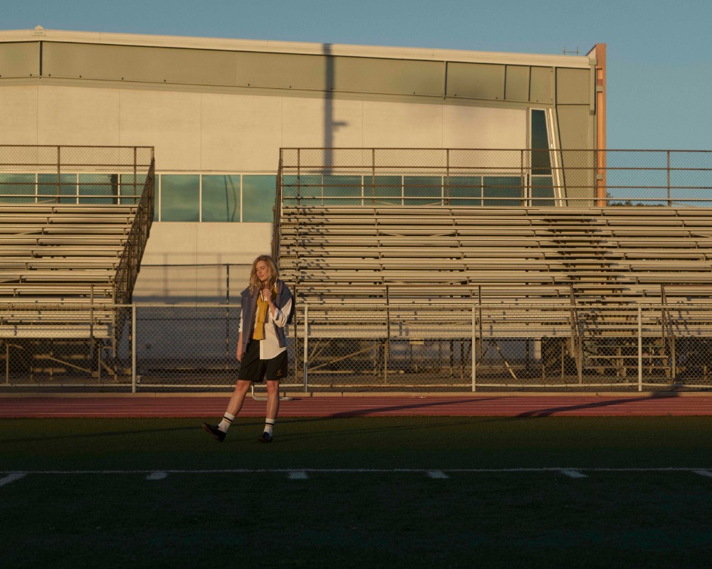 A woman stands on a sports field near stadium seating, wearing casual attire with socks and sneakers, under a clear blue sky at sunset.