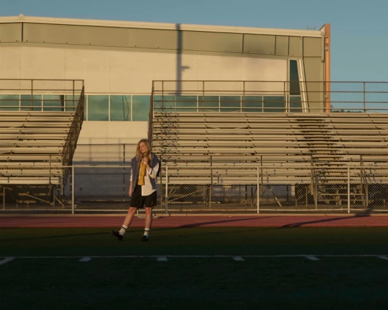 A woman stands on a sports field near stadium seating, wearing casual attire with socks and sneakers, under a clear blue sky at sunset.