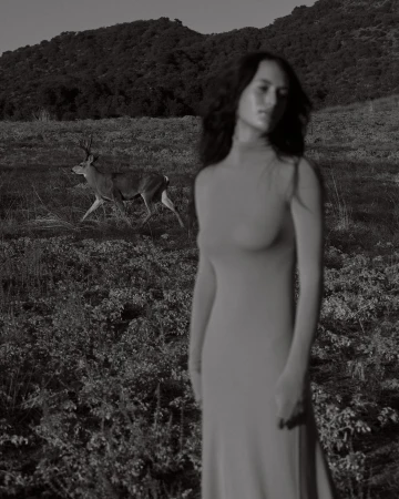 Monochrome portrait of a woman in a long dress standing in a field with a deer walking in the background.