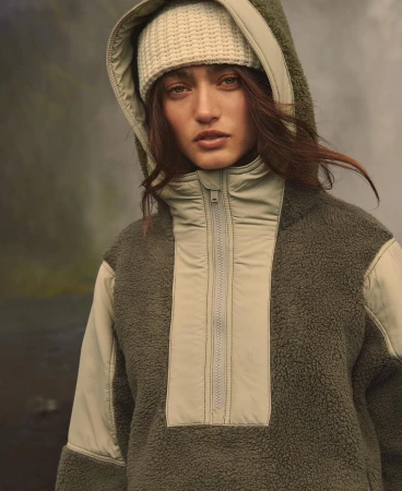 Close-up portrait of a woman wearing an olive green fleece hoodie with a hood up and a cream-colored knit beanie outdoors.