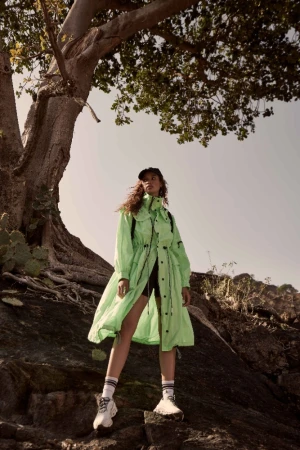 Model in neon green utility coat standing beneath a large tree on rocky terrain by Lindsey Childs