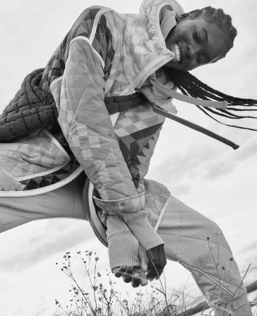 Black and white portrait of a smiling woman wearing a quilted patterned jacket outdoors under an overcast sky.