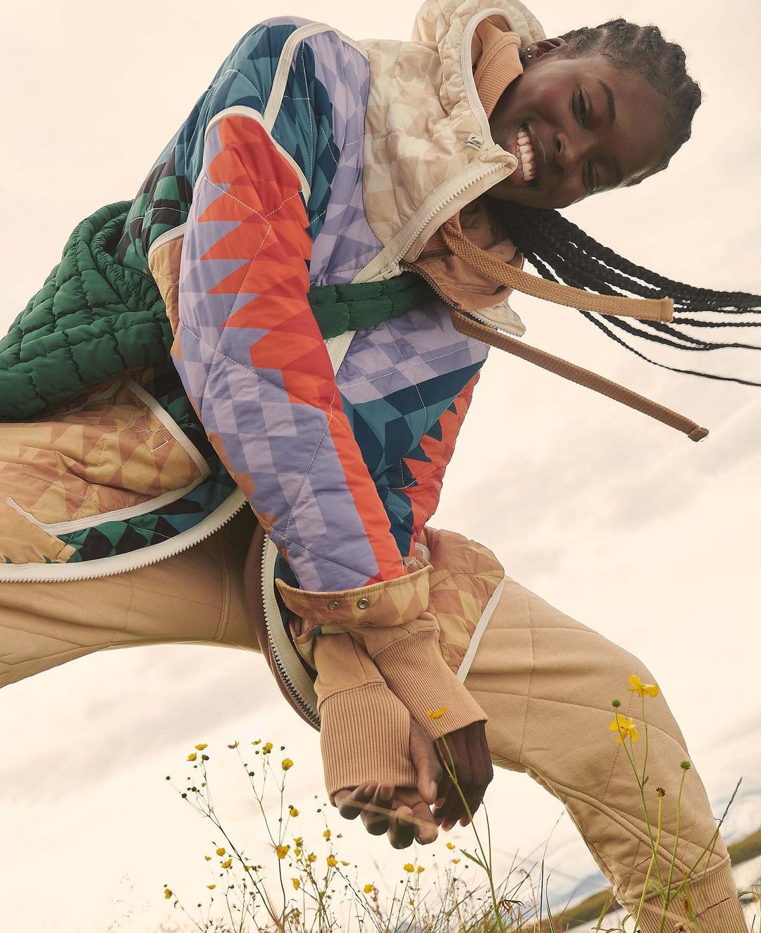 Smiling woman outdoors wearing a colorful quilted jacket and pants, looking up from a low angle with wild flowers and sky background.
