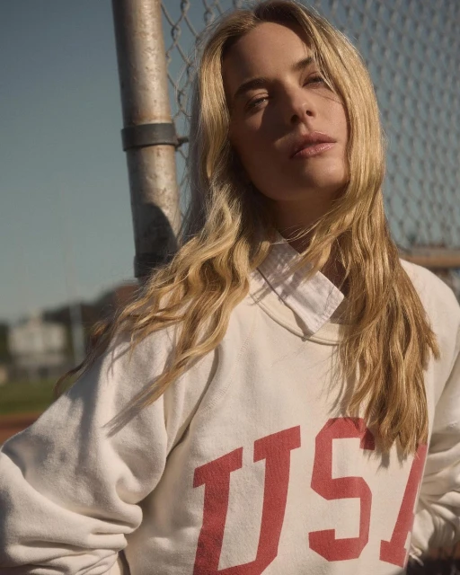 Portrait of a blonde woman wearing a sweatshirt with 'USA' lettering, posing in front of a chain-link fence on a sunny day.