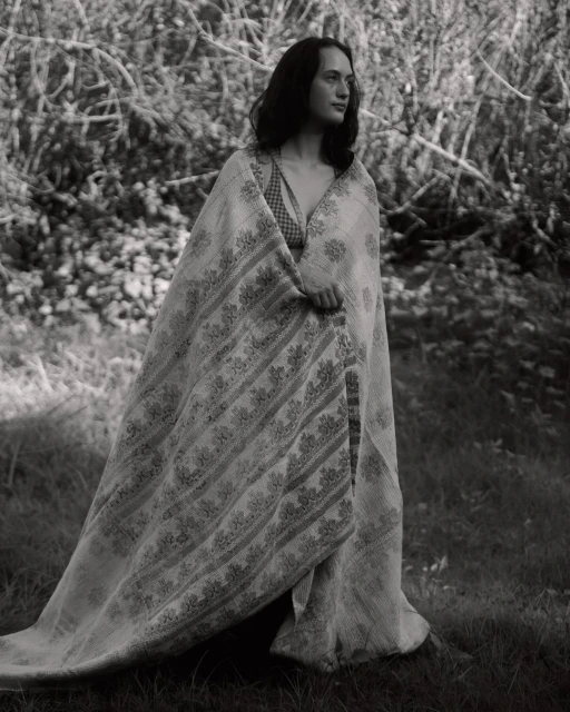 Monochrome portrait of a woman in a patterned shawl standing in a grassy field with blurred trees in the background, contemplative pose.