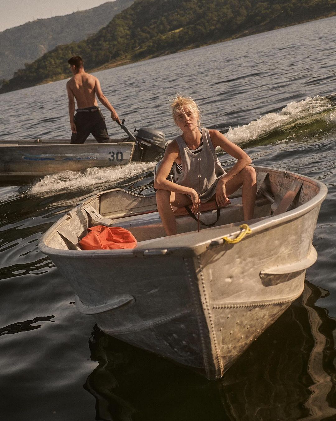 Woman sits in a rowboat on the water during daytime with a man in a rowboat in the background. Mountains and trees are visible.