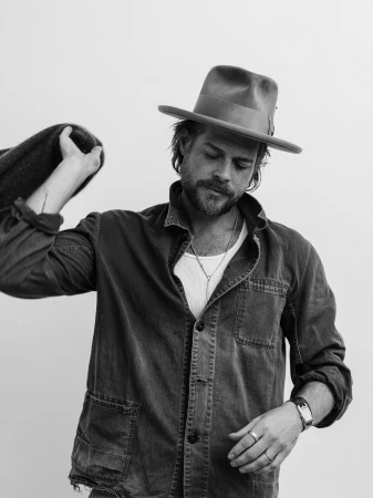 Man wearing a fedora and button-up shirt holding a jacket over his shoulder against a plain background.