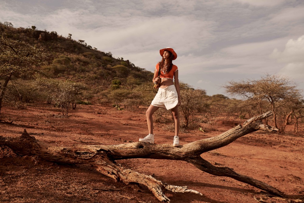 Model standing on a fallen tree branch in an arid landscape, wearing an orange top, white shorts, and a wide-brimmed hat.