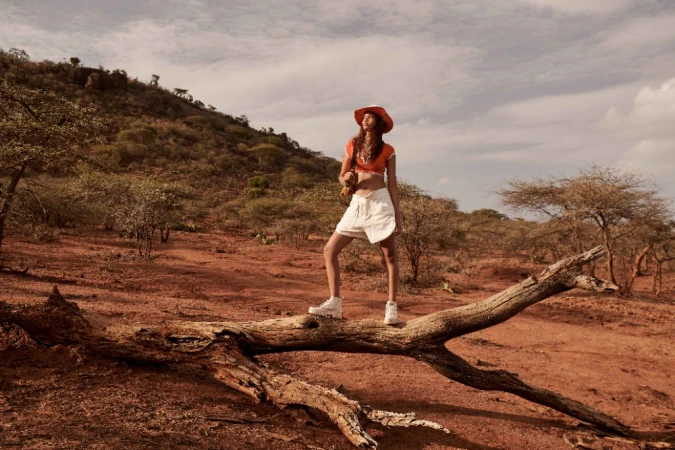 Model standing on a fallen tree branch in an arid landscape, wearing an orange top, white shorts, and a wide-brimmed hat.