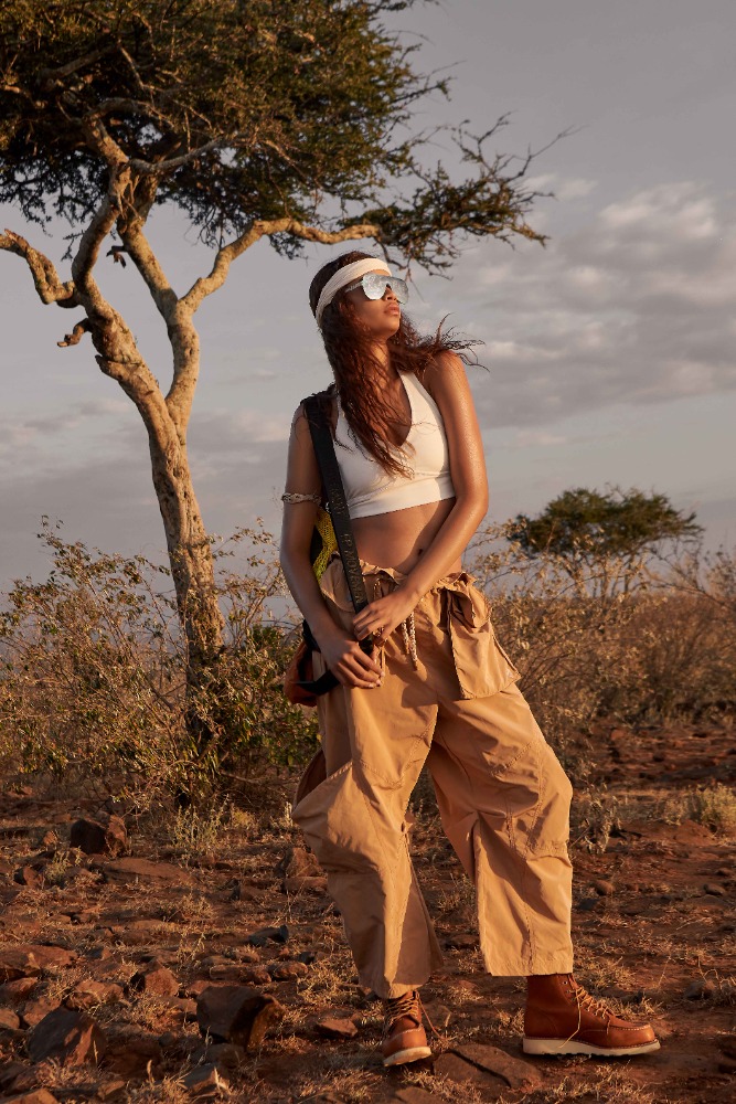 A woman in tan wide-leg pants, boots, and a white top stands in a savanna landscape near a tree, modelling in golden-hour sunlight.