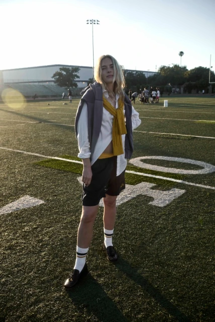 Woman in athletic shorts, socks, loafers, sweater draped over shoulder, posing on a football field, surrounded by a blurred group of people.