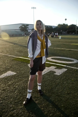 Woman in athletic shorts, socks, loafers, sweater draped over shoulder, posing on a football field, surrounded by a blurred group of people.