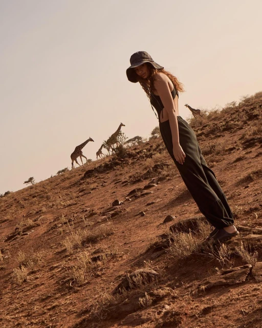 Woman in flowing pants and hat posing on a hillside in front of a group of giraffes in a natural outdoor environment.