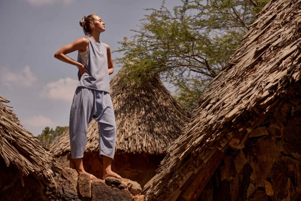 Adult female model wearing a blue linen tank top and matching pants poses on rocky terrain next to thatch-roofed buildings.