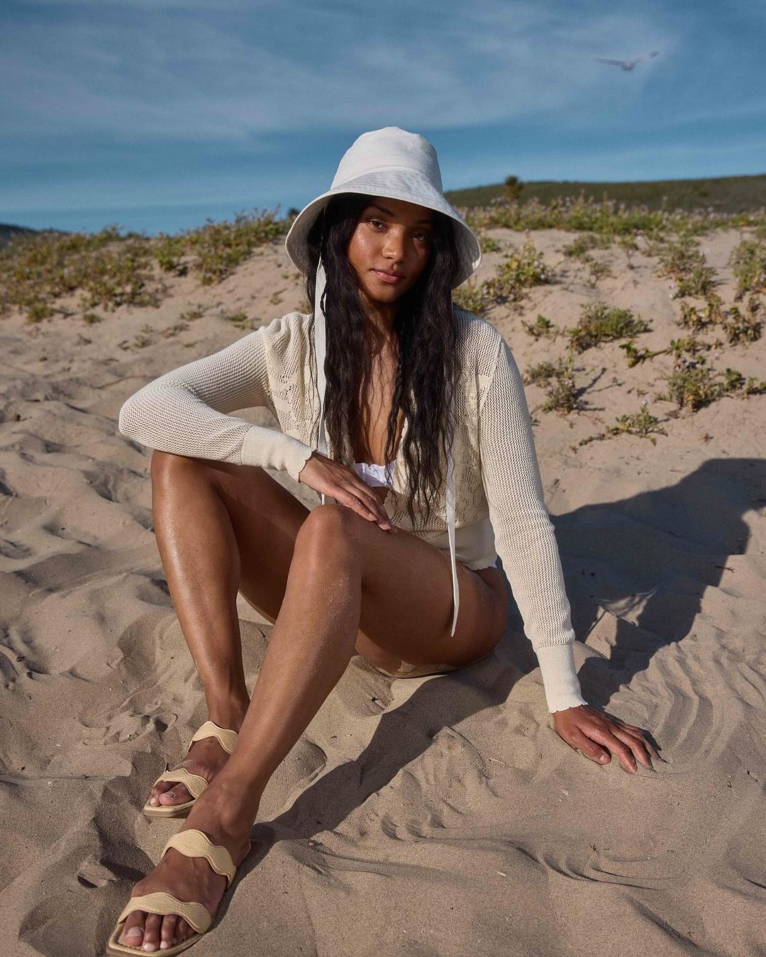A woman in a sun hat and cardigan sits on a sandy beach under a blue sky. She is wearing sandals. Coastal scene.