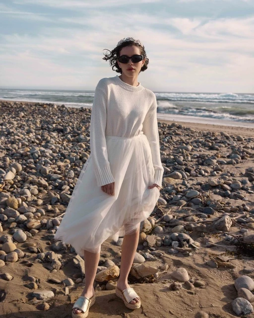 Woman in a white sweater and tulle skirt poses on a rocky beach under a bright sky with ocean waves in the background, wearing sunglasses.