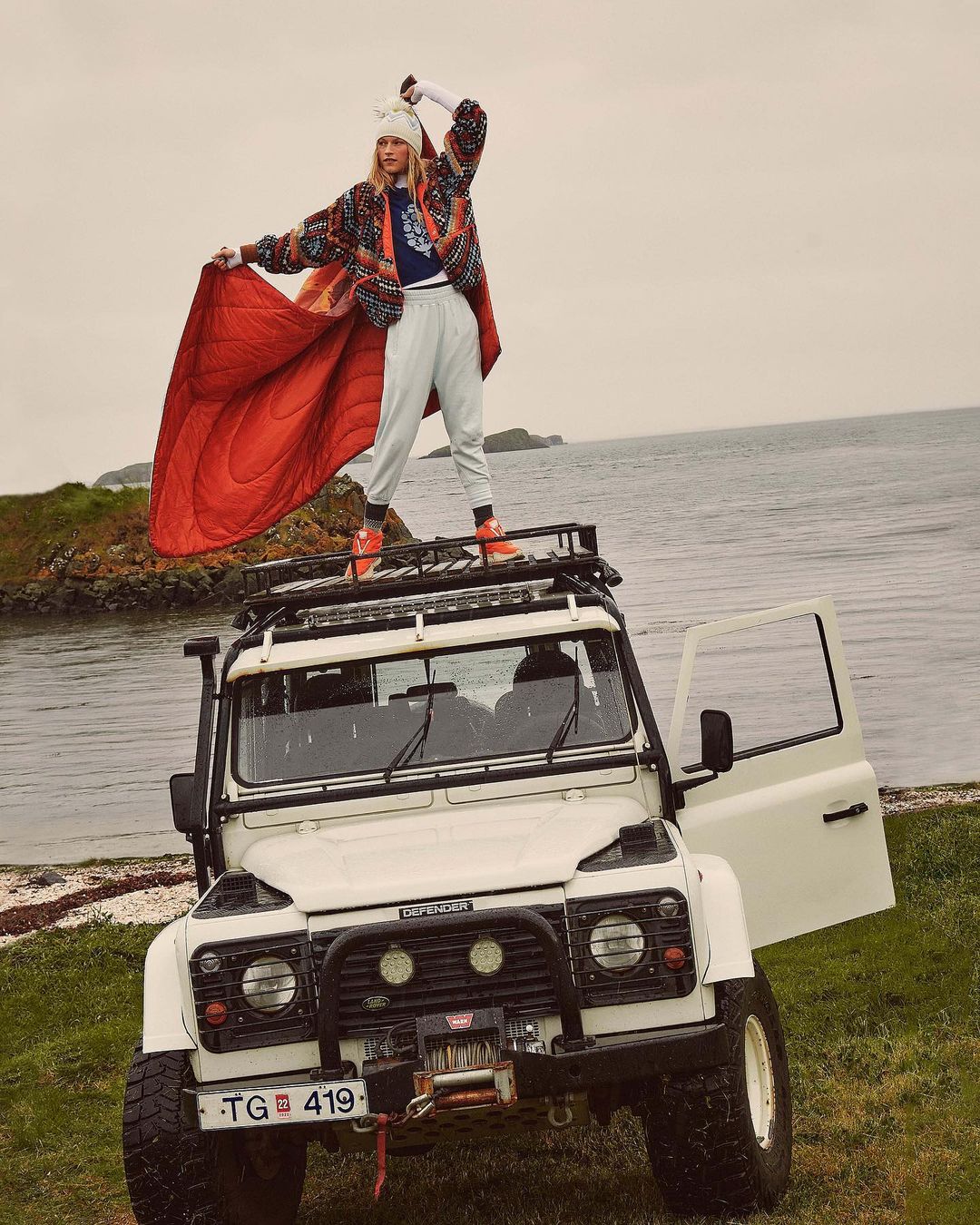 Young woman poses on a Land Rover Defender roof rack by the ocean wearing a patterned jacket, beanie, and sweatpants, holding a sleeping bag.
