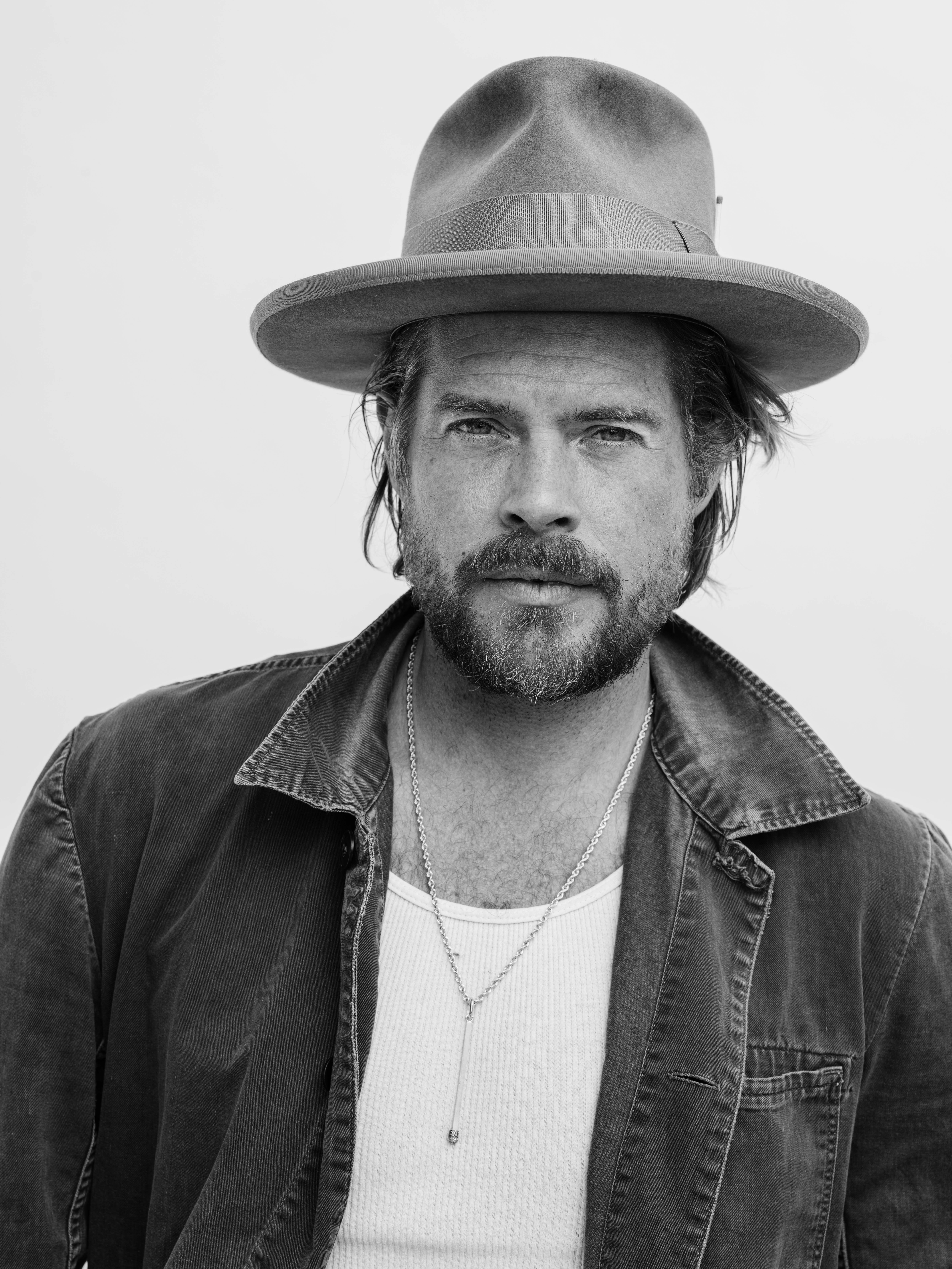Portrait of a bearded man wearing a wide-brimmed hat, tank top, and jacket against a plain background