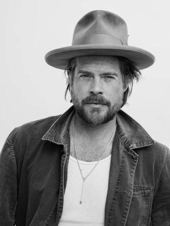 Portrait of a bearded man wearing a wide-brimmed hat, tank top, and jacket against a plain background