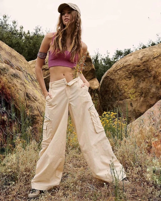 A slender adult woman poses outdoors wearing a mauve crop top, oversized tan trousers, baseball cap, and beaded arm cuff. 