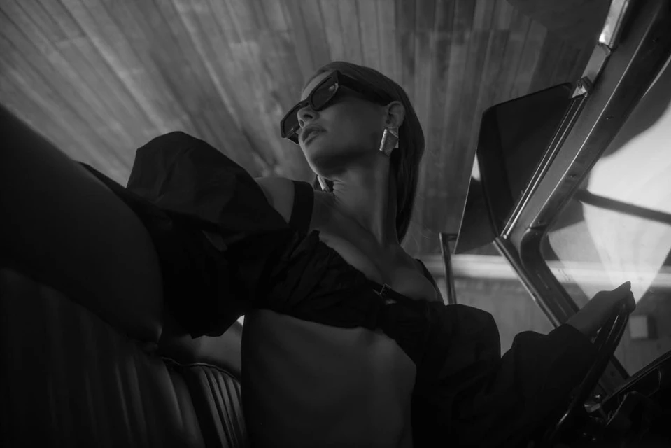 Monochrome low-angle shot of a woman in sunglasses behind the wheel of a vintage car, with wooden slats visible on the ceiling.