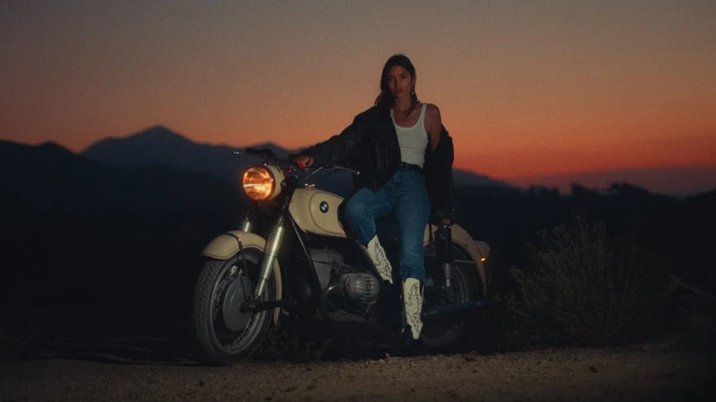 An adult female sits on a vintage BMW motorcycle in a desert landscape at dusk, wearing a tank top, jeans, cowboy boots, and a leather jacket.
