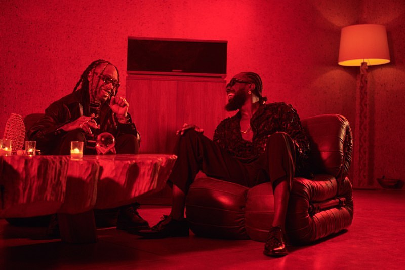 Two adult males sitting in a red-toned room. One man sits at a table with candles and a crystal ball, the other in a leather chair.