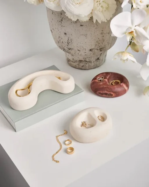 Close-up of cream and brown marbled jewelry holders with gold jewelry on a white table with a vase of white flowers in the background.