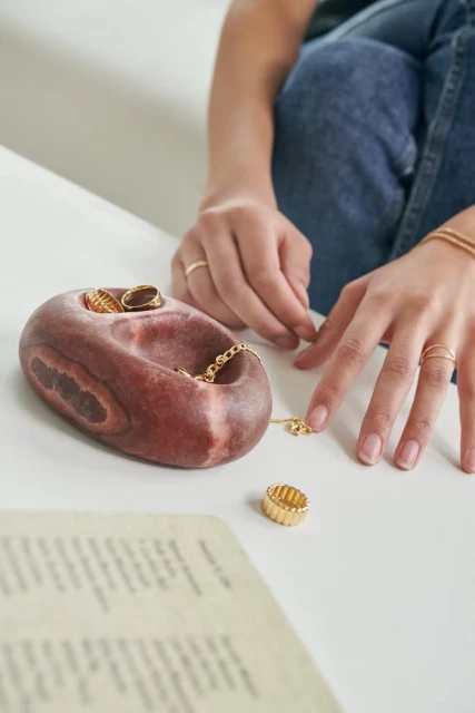 Close-up of a person arranging gold jewelry in a decorative stone bowl on a white table, with a focus on rings and a necklace.