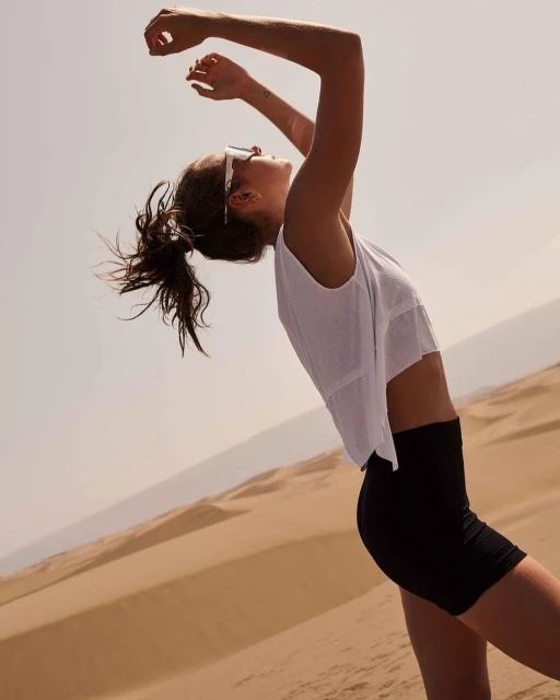Woman in white top and black shorts poses in a desert setting. The background includes sand dunes and a bright, clear sky.