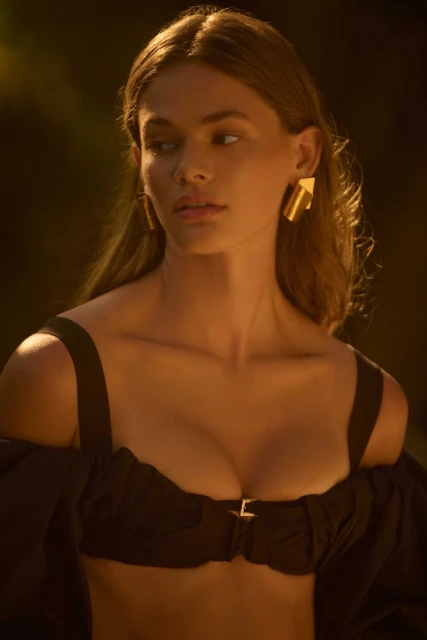 Woman in off-shoulder top with architectural gold earrings poses in a warm-toned outdoor shot with soft, diffused lighting.