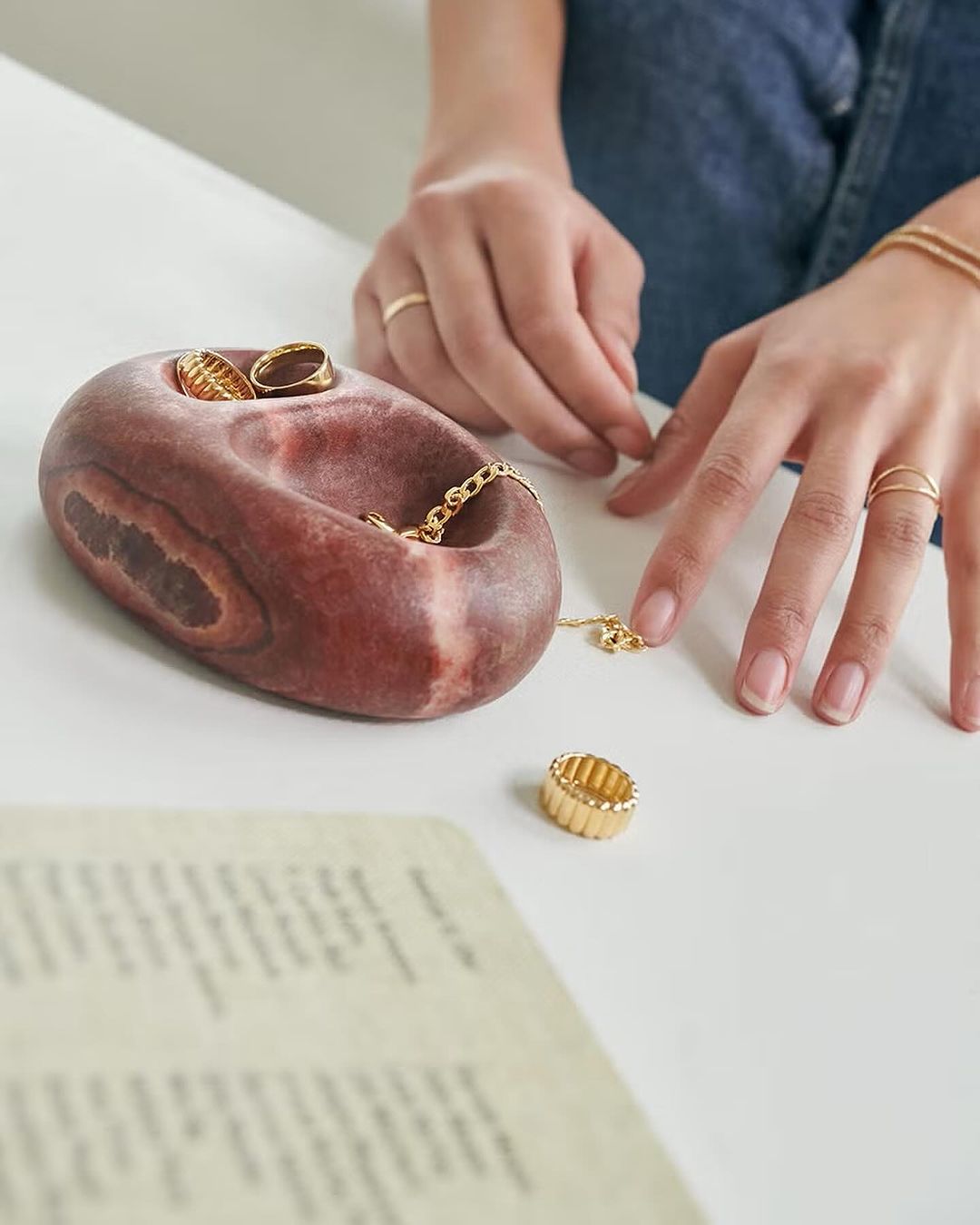 Close-up of a tan-skinned hand placing a gold ring next to a brown stone jewelry holder and other gold jewelry on a white surface.
