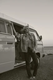 Blonde woman in denim jacket leaning against the open door of a vintage camper van by the coast.