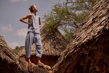A woman in a blue tank top and loose trousers stands on a stone wall between thatched-roof huts under an open sky.