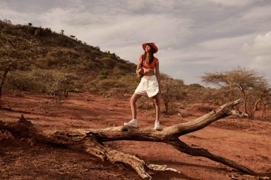 Model standing on a fallen tree branch in an arid landscape, wearing an orange top, white shorts, and a wide-brimmed hat.