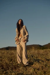 Woman wearing a beige matching jacket and trousers standing in a grassy meadow under a clear blue sky